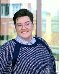 A butch presenting person smiling with glasses, a blue button up, and navy sweater in front of a window