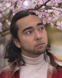 Photo of Alika Bourgette in a red shirt standing under a cherry tree with blossoms.