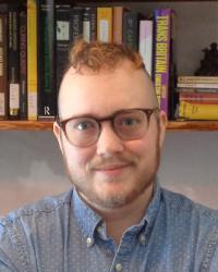 A person with short brown hair, a beard, and glasses, shown from the shoulders up. They are wearing a polka-dot blue shirt and are standing in front of a bookshelf.