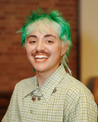 A mid-20s gender non conforming person with short green hair and a mustache. 