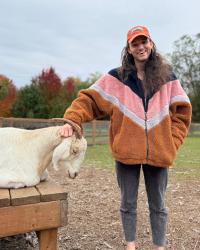 Photo of Jesse (a person with long hair, an orange hat, and a winter coat) smiling and petting a goat