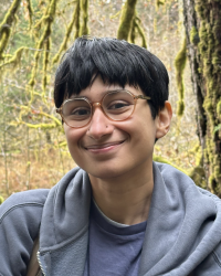 short haired brown person with glasses smiling with forest background