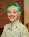 A mid-20s gender non conforming person with short green hair and a mustache. 